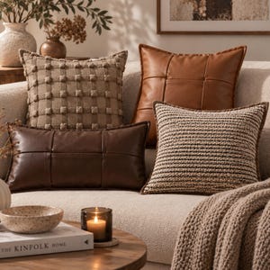 May include: A collection of four decorative throw pillows arranged on a sofa. One pillow features a tan grid pattern with raised tufts, one is a square tan faux leather with a grid stitch, one is a dark brown rectangular faux leather, and one is a textured knit.