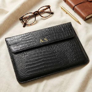 May include: Black faux-leather laptop sleeve with a crocodile skin texture. The sleeve has a flap closure and gold-colored initials. Tortoiseshell glasses, a gold pen, and a leather-bound notebook are also visible.