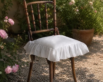 May include: A vintage wooden chair with a white, ruffled seat cushion. The chair has a spindle back and turned legs. The chair is set outdoors in a garden setting with pink roses and a terracotta pot in the background.