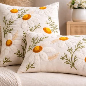 Può includere: Due cuscini decorativi bianchi con ricami a forma di fiori di margherita. Le margherite hanno centri gialli e petali bianchi, con foglie ricamate verdi e beige. I cuscini sono su una superficie bianca.