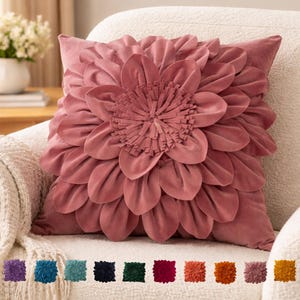 May include: A dusty rose-colored decorative pillow with a large, layered flower design. The flower's petals are soft and textured, with a fringed center. The pillow is square and sits on a cream-colored chair. A selection of colored pillows are shown below.