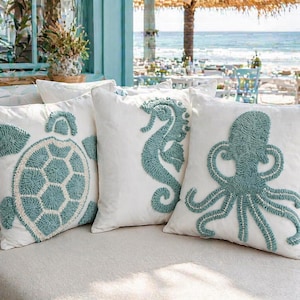 Puede incluir: Cuatro cojines blancos con diseños de criaturas marinas. Los cojines presentan una tortuga, un caballito de mar y un pulpo, todos en un diseño azul claro texturizado. Los cojines están dispuestos sobre una superficie de color claro, con una escena de playa al fondo.