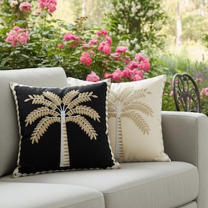 May include: Two decorative throw pillows with embroidered palm tree designs. One pillow is black with a gold palm tree, and the other is cream with a gold palm tree. Both pillows have a scalloped edge and are on a gray sofa.