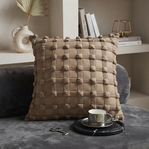 May include: A square throw pillow in a solid tan color featuring a grid pattern of raised, tufted pom-poms at each intersection. The pillow has a textured, woven appearance and is positioned on a dark gray surface with a metallic cup and a key.