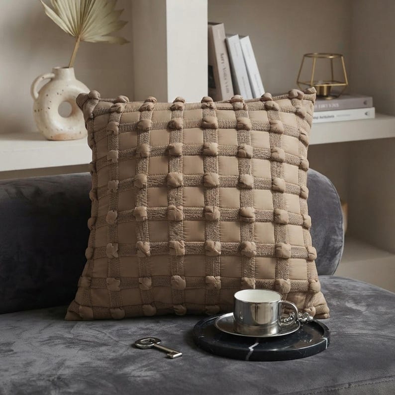 May include: A square throw pillow in a solid tan color featuring a grid pattern of raised, tufted pom-poms at each intersection. The pillow has a textured, woven appearance and is positioned on a dark gray surface with a metallic cup and a key.