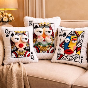 May include: Three playing card design cushions on a beige sofa. The cushions feature embroidered faces of a Queen, King, and Jack, with exaggerated features and bright colours. The cushions have a soft, white, furry texture.