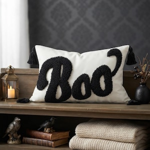 Puede incluir: Un cojín rectangular blanco que presenta la palabra Boo en letras grandes negras texturizadas hechas con técnica de punch needle. El cojín tiene borlas negras en cada una de las cuatro esquinas y un acabado de tela lisa en el cuerpo.