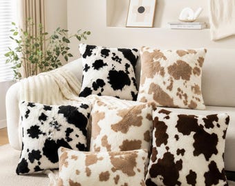May include: A collection of six decorative throw pillows with a cowhide pattern. The pillows feature a variety of colors, including black, brown, and tan, against a white background. The pillows are arranged on a white sofa.