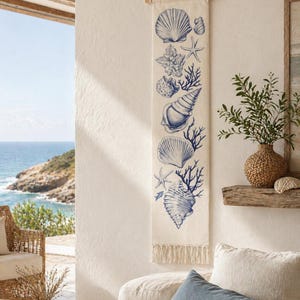 Op de afbeelding: Een crèmekleurige wandhanger met blauwe schelpenillustraties. Het ontwerp bevat verschillende schelpen, zeesterren en koraal, verticaal gerangschikt. De hanger is opgehangen aan een houten deuvel met een natuurlijk touw en heeft een gefranjerde onderkant.