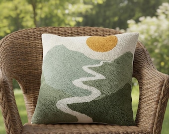May include: Square decorative pillow with a landscape design. The pillow features a textured surface with a mountain scene in shades of green, a yellow sun, and a white river. The pillow is placed on a wicker chair.