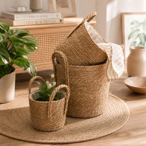 May include: A set of two cylindrical baskets crafted from thick, braided natural jute rope. The larger basket features a single handle and a light-colored fabric liner, while the smaller basket includes two handles and a green plant inside.
