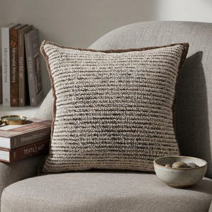 May include: A square throw pillow featuring a textured, horizontal knit pattern in shades of cream, beige, and dark brown. The pillow is finished with a solid brown fabric border along the edges, providing a structured frame to the soft, woven surface.