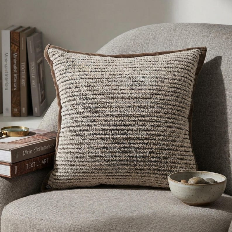 May include: A square throw pillow featuring a textured, horizontal knit pattern in shades of cream, beige, and dark brown. The pillow is finished with a solid brown fabric border along the edges, providing a structured frame to the soft, woven surface.
