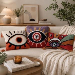 May include: Three square and rectangular throw pillows featuring tufted eye motifs. The left pillow is white with a large eye, the center is purple with a red and blue eye, and the right is a multi-colored sunburst design with a central eye.