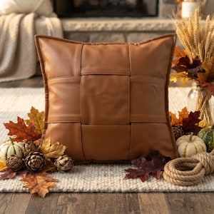 May include: A square throw pillow featuring a woven design made of smooth, tan-colored faux leather strips. The pillow has a structured, grid-like pattern created by the interlocking horizontal and vertical bands of material across the front surface.