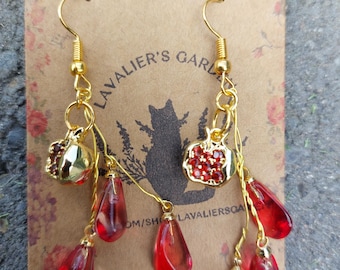 May include: Gold-tone dangle earrings featuring red teardrop-shaped beads and apple charms with red accents. The earrings hang from gold hooks. The background features a floral design and the text "Lavalier's Garden".