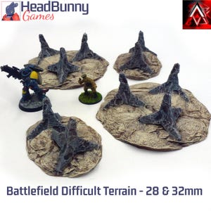 Peut inclure: Plusieurs éléments de terrain de champ de bataille miniatures avec des obstacles gris foncé sur des bases beiges. Deux figurines miniatures sont présentes. Le texte "Battlefield Difficult Terrain - 28 & 32mm" est en bas.