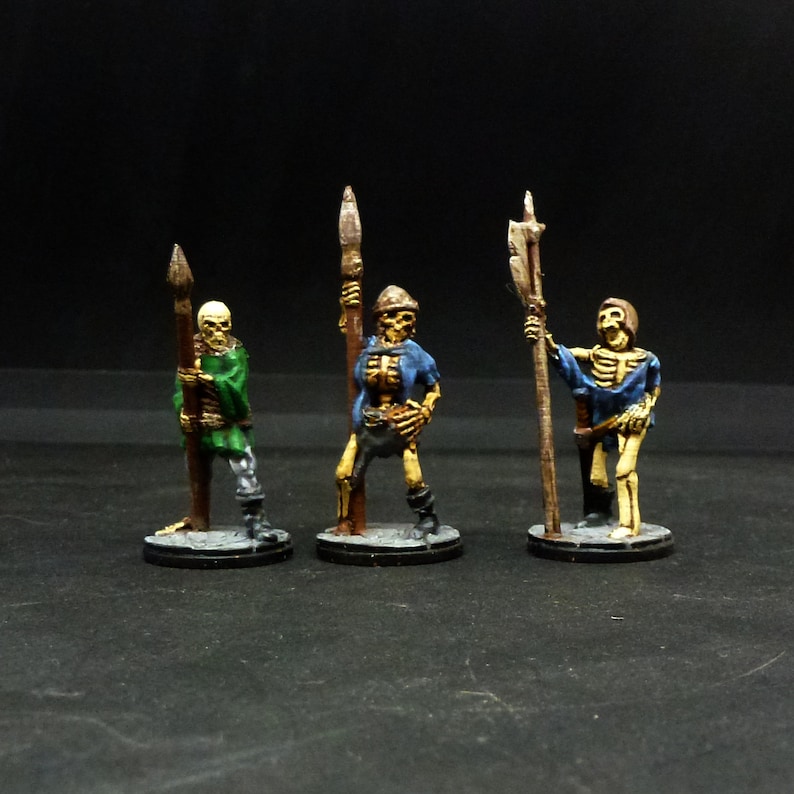 Peut inclure: Trois figurines squelettiques miniatures peintes tenant des lances, chacune v&ecirc;tue de v&ecirc;tements de couleurs diff&eacute;rentes. Les figurines sont debout sur des bases rondes.