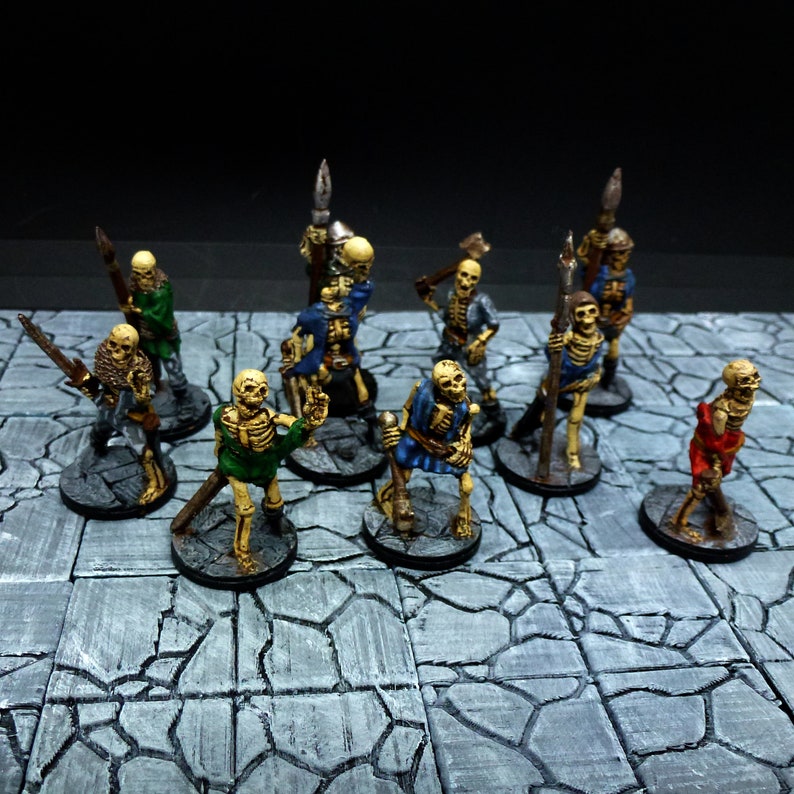 Peut inclure: Une collection de figurines squelettes miniatures peintes, certaines tenant des armes, debout sur une surface de pierre grise. Les squelettes sont peints dans diff&eacute;rentes couleurs, notamment le vert, le bleu et le rouge.