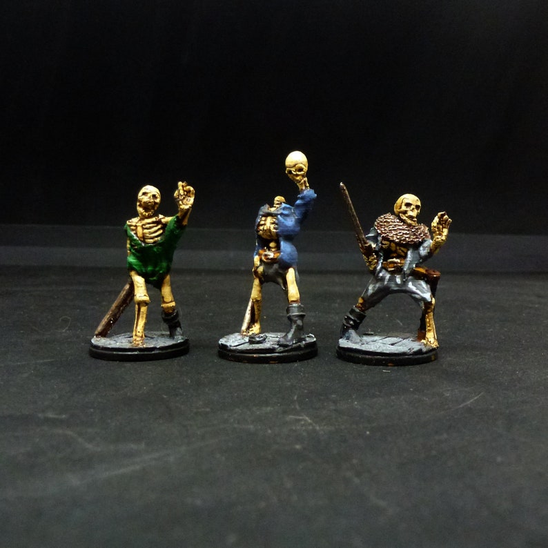 Peut inclure: Trois figurines squelettiques miniatures peintes, chacune avec une pose diff&eacute;rente et tenant un objet diff&eacute;rent. Les figurines sont debout sur des bases rondes.