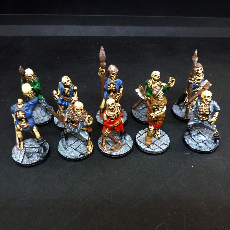 Peut inclure: Un groupe de dix figurines squelettes miniatures peintes, chacune avec une pose et une arme diff&eacute;rentes, debout sur des bases rondes. Les squelettes sont peints dans diff&eacute;rentes couleurs, notamment le bleu, le rouge, le vert et le brun.