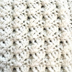 May include: Close-up of a white crocheted blanket. The textured surface features a repeating pattern of raised, flower-like stitches. The soft, off-white color and intricate design suggest a handmade item, perfect for home decor or as a cozy accessory.