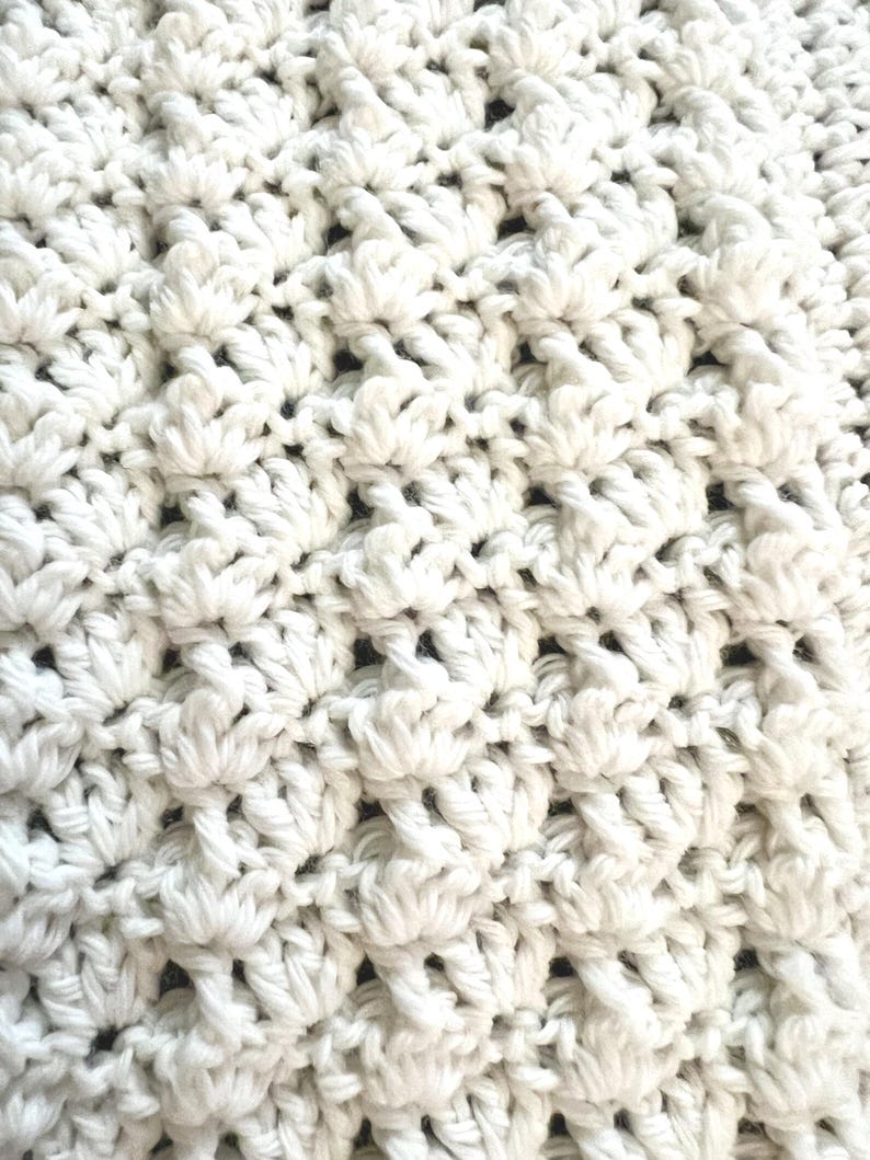 May include: Close-up of a white crocheted blanket. The textured surface features a repeating pattern of raised, flower-like stitches. The soft, off-white color and intricate design suggest a handmade item, perfect for home decor or as a cozy accessory.
