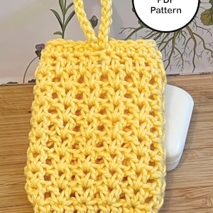 Puede incluir: Un jabonera de ganchillo de color amarillo brillante con un lazo para colgar. La jabonera es rectangular y tiene un diseño de tejido abierto y texturizado. El texto "Crochet PDF Pattern" está en un círculo blanco.