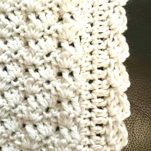 May include: Close-up of a white, textured crochet fabric. The crochet pattern features a dense, raised design with a scalloped edge. The fabric appears soft and handmade, with a focus on texture and detail.