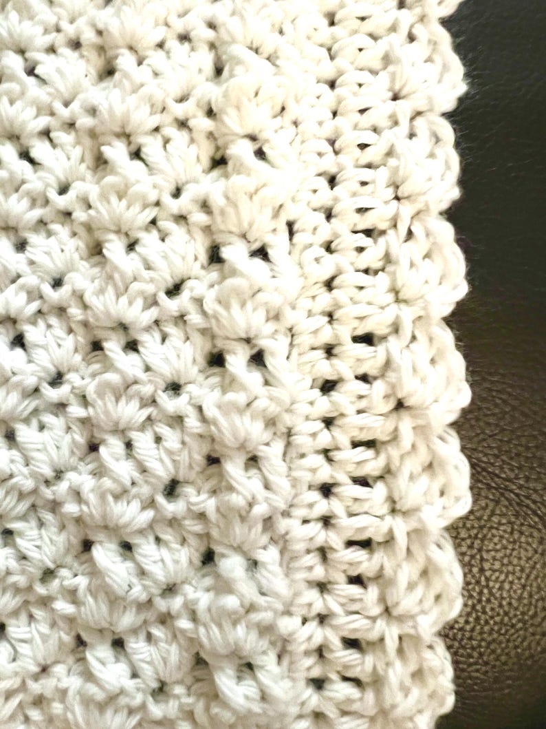 May include: Close-up of a white, textured crochet fabric. The crochet pattern features a dense, raised design with a scalloped edge. The fabric appears soft and handmade, with a focus on texture and detail.