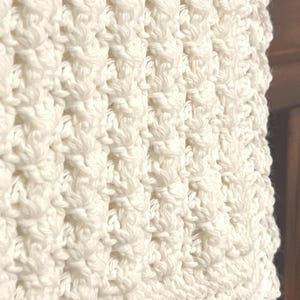 May include: Close-up of a cream-colored, textured crochet fabric. The pattern features a series of raised, textured stitches, creating a visually interesting surface. The crochet work appears to be tightly woven, with a soft, inviting appearance. The overall aesthetic is cozy and handmade.