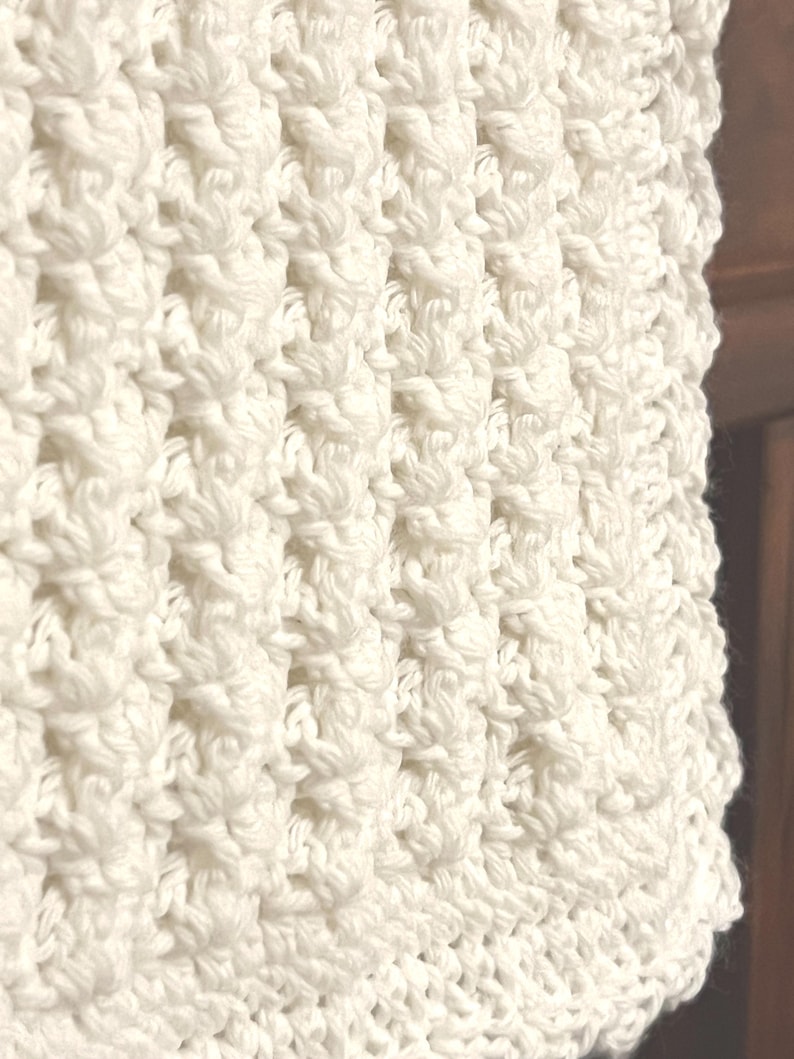 May include: Close-up of a cream-colored, textured crochet fabric. The pattern features a series of raised, textured stitches, creating a visually interesting surface. The crochet work appears to be tightly woven, with a soft, inviting appearance. The overall aesthetic is cozy and handmade.