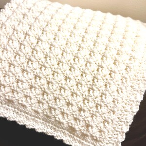 May include: A folded, off-white crochet blanket. The blanket has a textured pattern and a scalloped edge. The soft, handmade blanket is likely made of a natural fiber, such as cotton or wool. The blanket is folded on a dark surface.