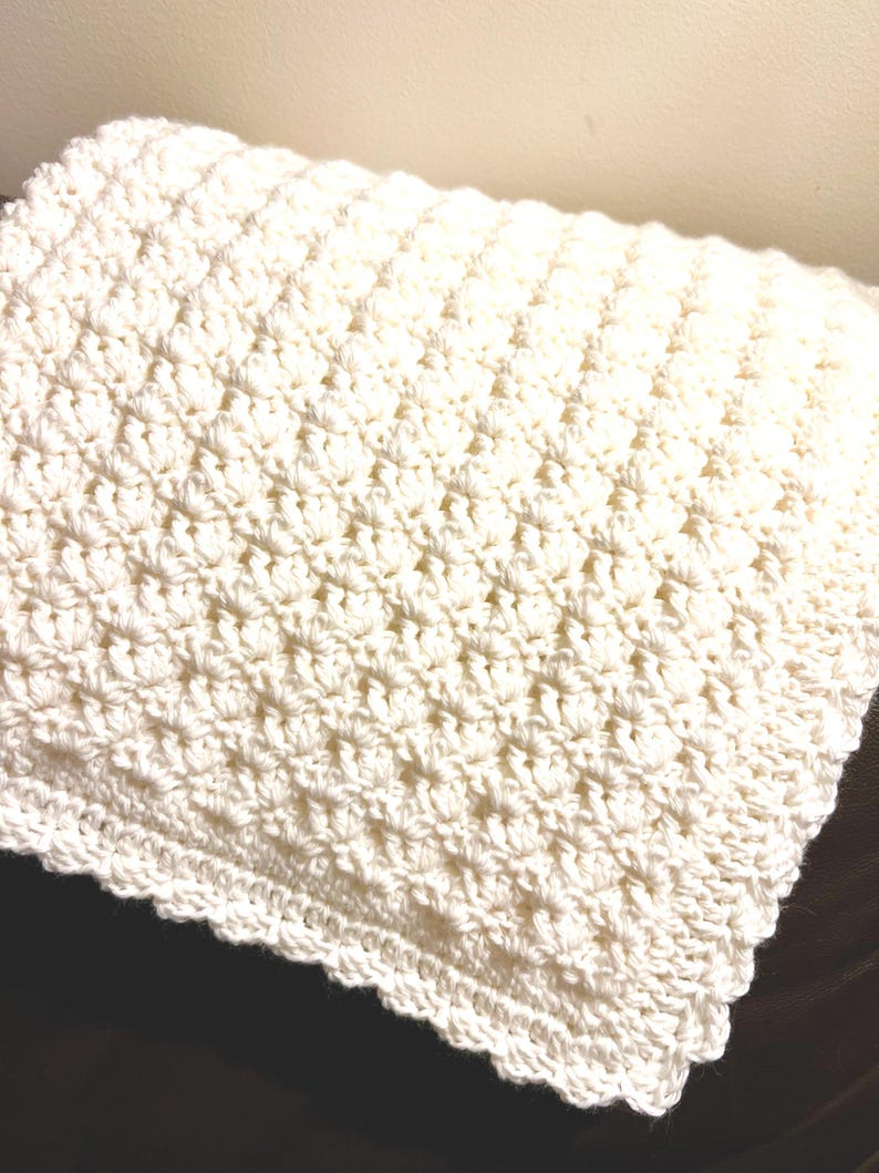 May include: A folded, off-white crochet blanket. The blanket has a textured pattern and a scalloped edge. The soft, handmade blanket is likely made of a natural fiber, such as cotton or wool. The blanket is folded on a dark surface.