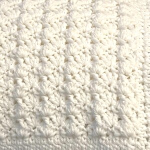 May include: Close-up of a white crocheted blanket. The blanket features a textured pattern with raised, flower-like designs. The crochet work is dense and detailed, with a visible stitch pattern. The overall appearance is soft and inviting, suggesting a handmade item.