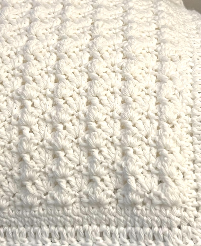 May include: Close-up of a white crocheted blanket. The blanket features a textured pattern with raised, flower-like designs. The crochet work is dense and detailed, with a visible stitch pattern. The overall appearance is soft and inviting, suggesting a handmade item.