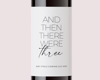 May include: A dark glass wine bottle featuring a white rectangular label with black text that reads AND THEN THERE WERE three, followed by the smaller text BABY STEELE COMING JULY 2025. The bottle has a tall neck and a dark grey foil cap.