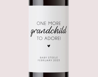 May include: A wine bottle with a white label that says "One more grandchild to adore!" with a small heart symbol and the text "Baby Steele, February 2023".