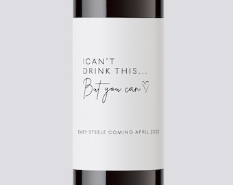 May include: A wine bottle with a white label that reads "I can't drink this... But you can." with a heart symbol. The label also says "Baby Steele coming April 2022".