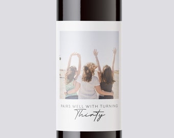 May include: A wine bottle with a white label featuring a photo of three people with their arms raised in the air. The label also includes the text "Pairs well with turning Thirty."
