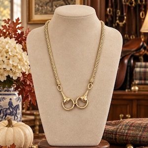 May include: A gold-toned necklace featuring a textured chain and a central horse bit pendant. The piece consists of two circular rings connected by a horizontal bar, mimicking equestrian hardware, displayed on a neutral-colored jewelry bust.