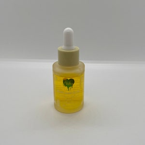 May include: A clear glass bottle containing a yellow liquid. The bottle has a white dropper top and a green heart-shaped sticker with the text "Just Kaya" on it. The bottle is labelled "Face Serum".
