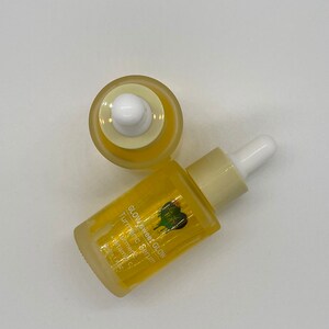 May include: A yellow glass bottle with a dropper top filled with a yellow liquid. The bottle is labelled "Glow Sweet Glow Turmeric Serum" with a green heart logo. The bottle is on a white surface.