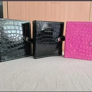 Op de afbeelding: Drie vierkante organizers met een glanzende krokodil-reliëftextuur. Twee organizers zijn zwart en één is felroze. Elke organizer heeft een zijband met een zilverkleurige metalen drukknoopsluiting en zichtbare witte stiksels langs de randen.