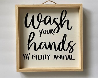 May include: A wooden framed sign with the text "Wash your hands ya' filthy animal" in black cursive lettering on a white background.