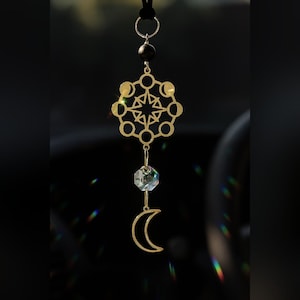 May include: A gold-colored celestial suncatcher with a black cord. The design includes a starburst, moon phases, a crystal, and a crescent moon. The suncatcher is suspended against a dark background, with light refracting through the crystal.