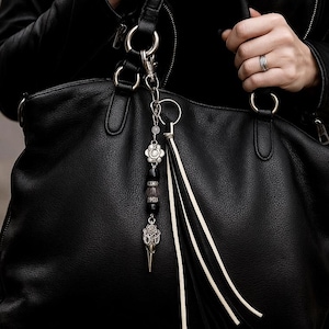 May include: Black leather handbag with a decorative charm. The charm features a silver-toned flower, beads, and a bird skull pendant. The bag has silver hardware and a tassel. The person is wearing a black leather jacket and rings.