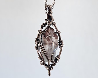 May include: A handcrafted pendant necklace featuring a clear quartz crystal encased in an ornate, wire-wrapped setting. The pendant hangs from a delicate chain with small, decorative beads. The wirework is a dark, antique copper colour.