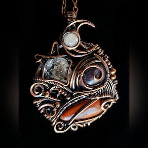 Puede incluir: Collar con colgante envuelto en alambre de cobre con un diseño único. El colgante presenta una piedra transparente en bruto, una piedra de color púrpura oscuro y una piedra marrón anaranjada. Una piedra blanca y redonda está colocada en la parte superior. El collar cuelga de una cadena de cobre.