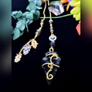 May include: A handmade, gold-toned charm with a black arrowhead-shaped stone wrapped in gold wire. The charm is suspended from a gold chain with black beads, an evil eye charm, and skull, owl, and hand charms. The background is black.